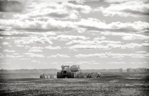 A tractor pulling a seeder and fertilizer tanks planting a crop in a black and white countryside summer landscape